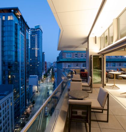 The image shows a modern urban balcony at twilight, complete with seating, tables, and a view of high-rise buildings and a city street below.