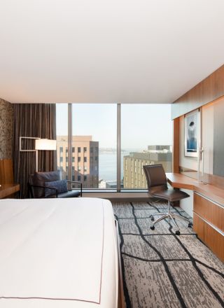 A modern hotel room with a large bed, wall-mounted TV, desk area, chair, and a window with a cityscape view.