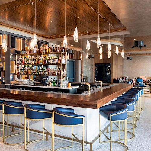 A modern bar with a variety of bottles, blue barstools, pendant lights, and a spacious seating area in the background.