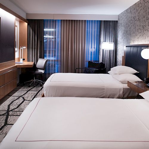 A modern hotel room with two double beds, a TV on the wall, a desk with a chair by the window, and contemporary decor.