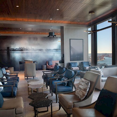 A modern lounge with comfy chairs, large windows, and a cityscape mural on the back wall, offering a scenic waterfront view and a Ferris wheel.