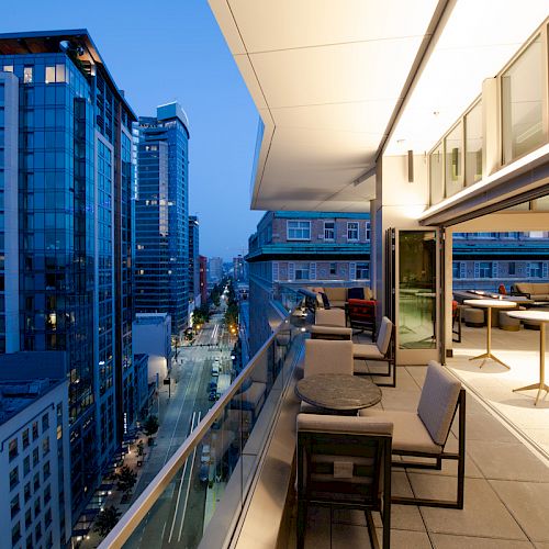 This image shows a modern cityscape at dusk with a view from a high-rise balcony with seating and tables, overlooking a street lined with tall buildings.