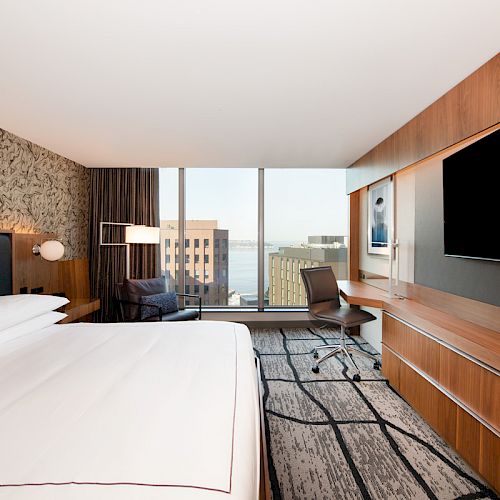 The image shows a modern hotel room with a large bed, a desk with a chair, a flat-screen TV, and a window with a city view.