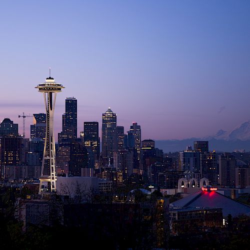The image shows the skyline of Seattle at dusk, with the Space Needle prominently featured, city buildings, and Mount Rainier in the background.