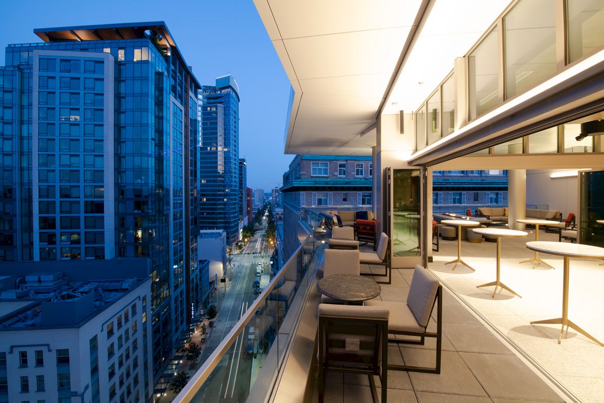 A modern balcony overlooks a cityscape with tall buildings and a street below, featuring tables, chairs, and a well-lit interior.