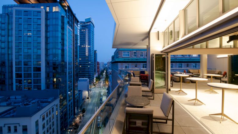 A modern balcony overlooks a cityscape with tall buildings and a street below, featuring tables, chairs, and a well-lit interior.