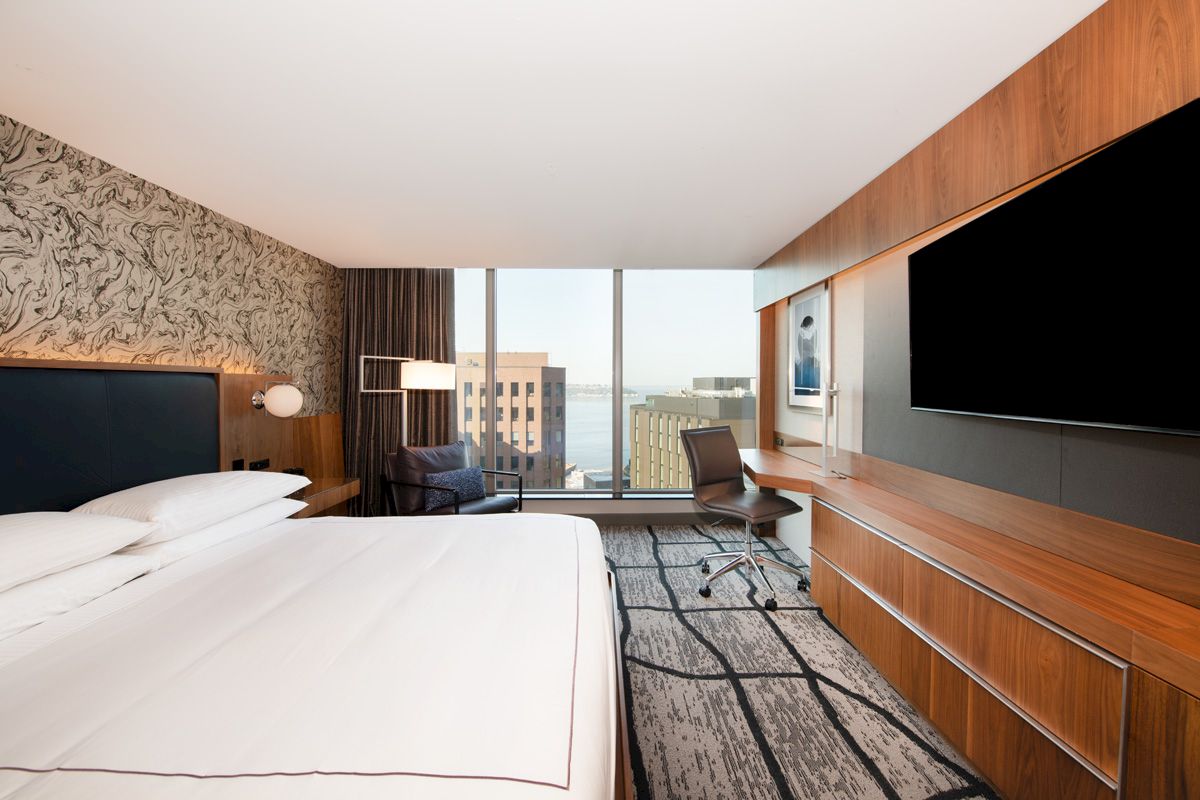 A modern hotel room with a large bed, abstract wallpaper, a desk with a chair, a TV, and a view of city buildings through a large window.