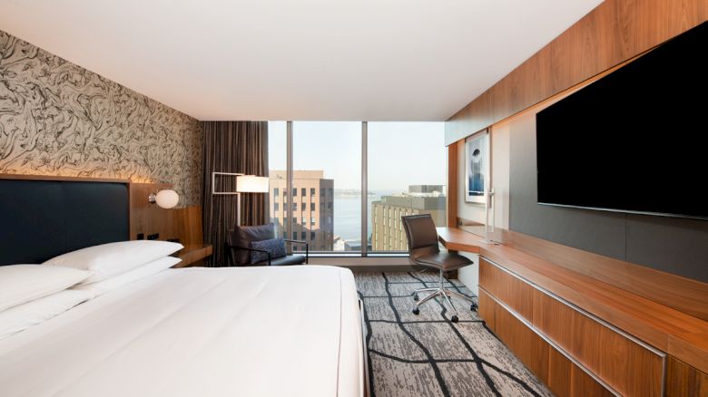 A modern hotel room with a large bed, abstract wallpaper, a desk with a chair, a TV, and a view of city buildings through a large window.