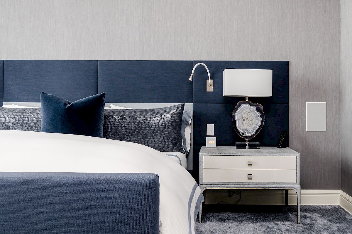 A modern bedroom with a navy blue headboard, white lamp, and nightstand placed on a grey carpet, featuring a neatly made bed with blue and white linens.