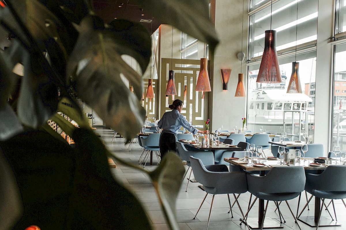 A person is setting tables in a stylish restaurant with modern decor and large windows, featuring teal chairs and orange pendant lights.