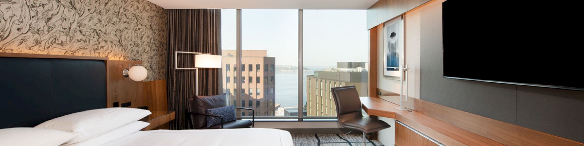 A modern hotel room with a large bed, a wall-mounted TV, a desk, and a chair near a window offering a city view with buildings in the background.